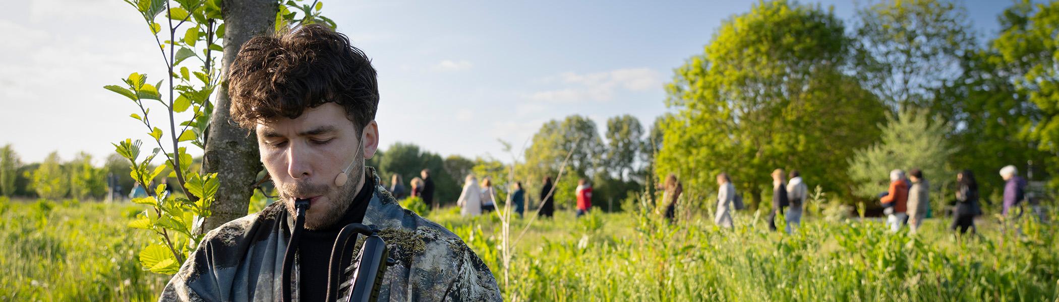 man bespeelt fluit op de achtergrond een weiland met hoog gras en koeien